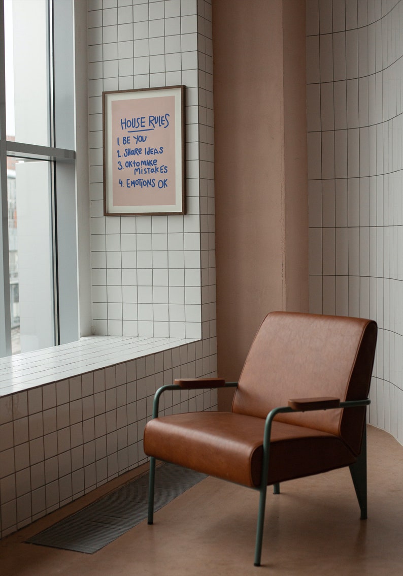 Pu&ograve; includere: Una stampa incorniciata con il testo "HOUSE RULES" e le frasi "BE YOU", "SHARE IDEAS", "OK to make MISTAKES" e "EMOTIONS OK". Una poltrona in pelle marrone con struttura in metallo verde &egrave; di fronte a un muro piastrellato.