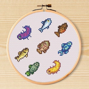 May include: A cross-stitch embroidery hoop with a white background and eight different coloured fish designs. The fish are in various shapes and sizes, including a purple fish, a blue fish, a yellow fish, a brown fish, and a green fish.