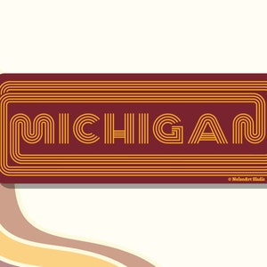 May include: A burgundy rectangular sticker with the word "MICHIGAN" in a retro, layered, gold font. The sticker has a subtle shadow effect.