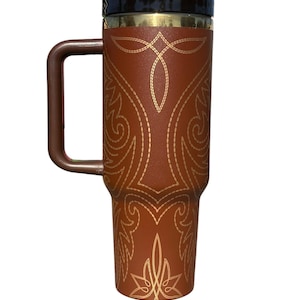 May include: A brown travel mug with a handle and a black lid. The mug features a gold decorative pattern. The handle and lid are brown. The mug is designed to keep beverages hot or cold.