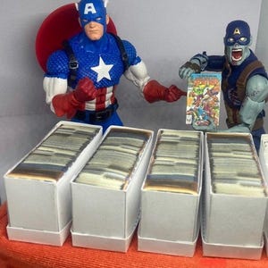 May include: Five white boxes filled with trading cards. The boxes are arranged in a row. A red, white, and blue action figure of Captain America stands to the left of the boxes. A zombie action figure of Captain America holds a comic book in front of the boxes.