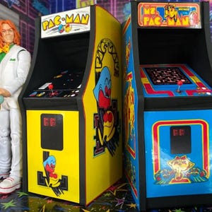 May include: Two miniature arcade game machines, one yellow and one blue, featuring Pac-Man and Ms. Pac-Man graphics. The yellow machine has a Pac-Man character on the side. The blue machine has a Ms. Pac-Man character. Both have game screens and joysticks.
