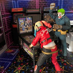 May include: Two action figures in an arcade setting. One figure with blonde and pink hair wears a red and black jacket and pants, leaning over an Elvira pinball machine. The other figure has green hair and a white mask, wearing a dark blue jumpsuit.
