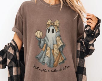 May include: Brown t-shirt featuring a cartoon ghost holding a baseball and a bat. The ghost is wearing a patchwork outfit and a bow. The text "Fall nights & ballpark lights" is printed below the graphic.