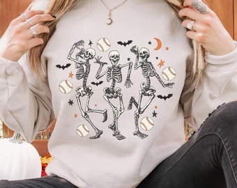 May include: Cream-colored sweatshirt featuring a graphic of three dancing skeletons with baseballs, bats, stars, and a crescent moon. The design is in black and orange. A casual, long-sleeved top for Halloween or sports fans.