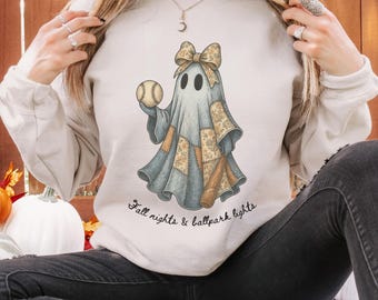 May include: Cream-colored sweatshirt featuring a ghost holding a baseball. The ghost is adorned with a bow and patchwork details in shades of blue, tan, and brown. The text "Fall nights & ballpark lights" is written in a script font.