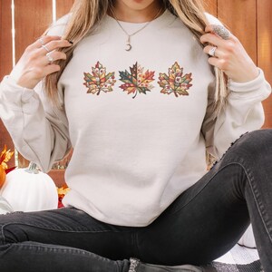 May include: A cream-colored sweatshirt featuring three embroidered autumn leaves in shades of red, orange, and green. The leaves have intricate floral patterns within their outlines. The person is wearing black jeans.