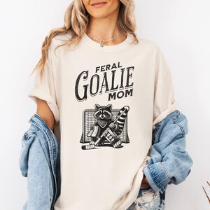 May include: A cream-colored t-shirt with the text "Feral Goalie Mom" in black lettering. A black and white illustration of a raccoon wearing hockey gear and standing in front of a hockey net is below the text.