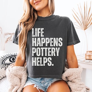 May include: A woman wearing a dark gray cropped t-shirt with the text "Life Happens Pottery Helps." in white lettering. She is wearing denim shorts and a white fuzzy jacket.