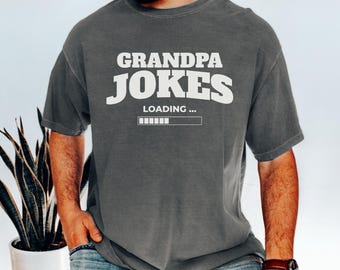 May include: A gray t-shirt with the words "GRANDPA JOKES" in white block letters, with "LOADING..." and a loading bar graphic underneath. The shirt is short-sleeved and has a relaxed fit.