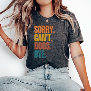 May include: A dark gray t-shirt with the text "SORRY. CAN'T. DOGS. BYE." in a vintage style font with colorful letters. The letters are in shades of orange, yellow, blue, and green.