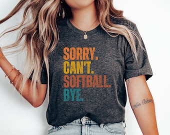 May include: A dark gray t-shirt with the text "SORRY. CAN'T. SOFTBALL. BYE." in a retro font with orange, yellow, blue, and green colors.