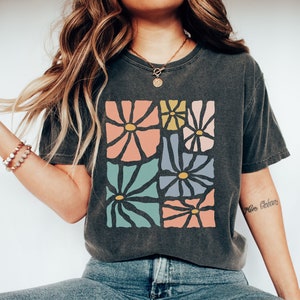 Op de afbeelding: Een donkergrijs T-shirt met een kleurrijk abstract bloemenontwerp. Het ontwerp bevat verschillende bloemen in tinten roze, blauw, groen en geel.