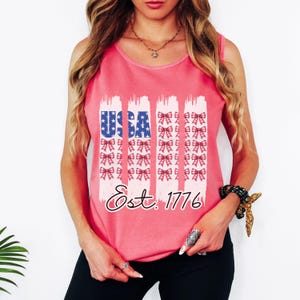 May include: A pink vest top featuring a patriotic design. The word "USA" is in blue with white stars, and the text "Est. 1776" is below. Vertical white brushstrokes with red bows are on the top.
