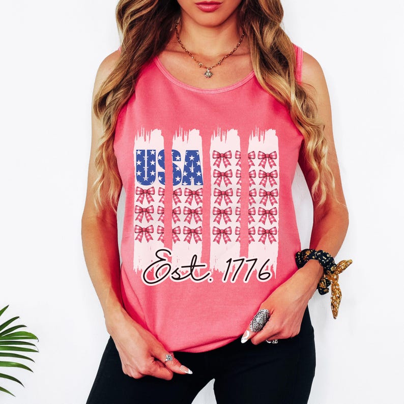 May include: A pink vest top featuring a patriotic design. The word "USA" is in blue with white stars, and the text "Est. 1776" is below. Vertical white brushstrokes with red bows are on the top.