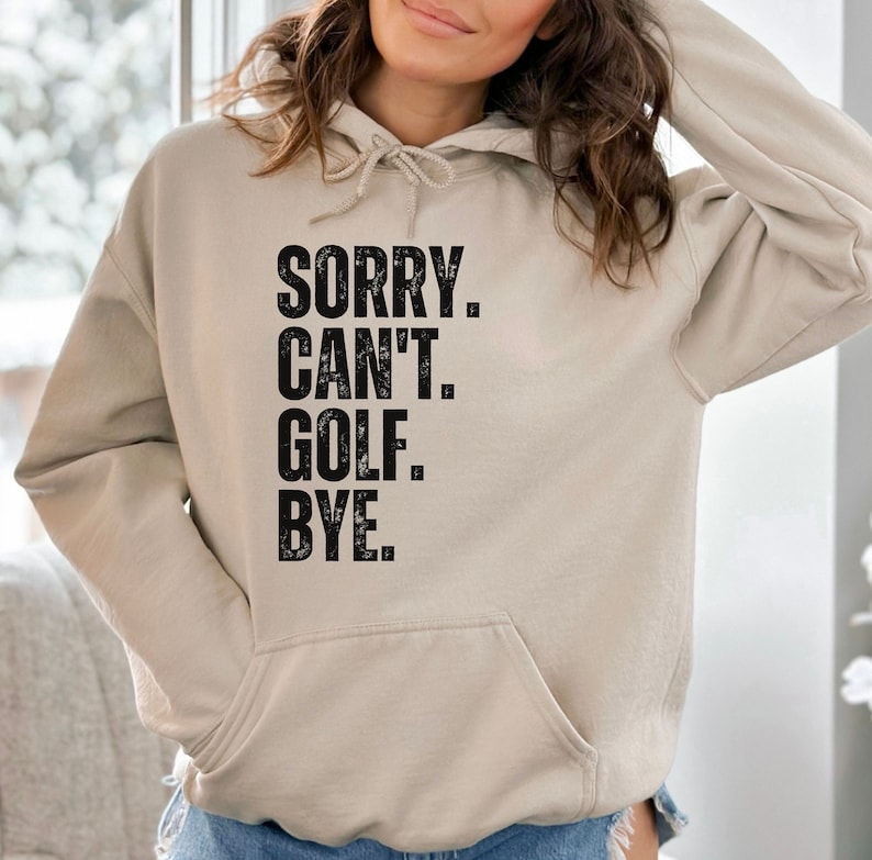 May include: A beige hoodie with a black print that says "SORRY. CAN'T. GOLF. BYE."