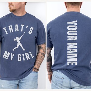 May include: A navy blue t-shirt with a white graphic of a girl playing baseball and the text "That's My Girl". The back of the shirt has the text "Your Name" in white.
