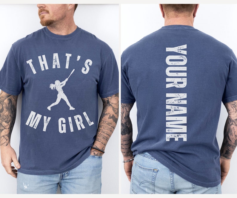 May include: A navy blue t-shirt with a white graphic of a girl playing baseball and the text "That's My Girl". The back of the shirt has the text "Your Name" in white.