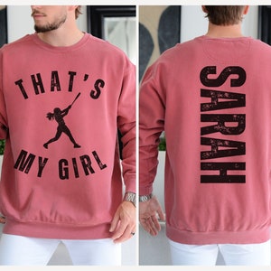 May include: A red sweatshirt with a black graphic on the front that says "That's My Girl" with a silhouette of a girl playing baseball. The back of the sweatshirt has the name "SARAH" printed in black.