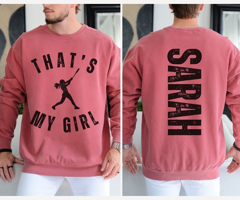 May include: A red sweatshirt with a black graphic on the front that says "That's My Girl" with a silhouette of a girl playing baseball. The back of the sweatshirt has the name "SARAH" printed in black.