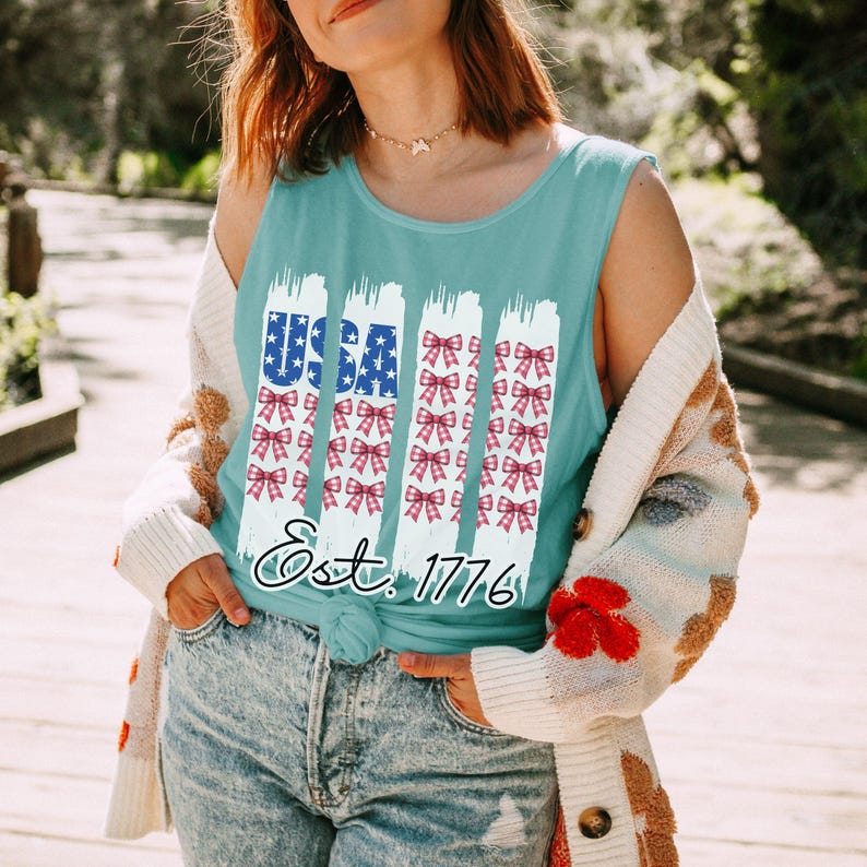 May include: A turquoise vest top featuring a patriotic design. The word "USA" is in blue and white, with red and white bows below. The text "Est. 1776" is in a cursive font. Worn with blue jeans and a patterned cardigan.