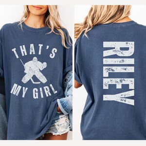 May include: A blue t-shirt with a white graphic of a hockey player and the text "That's My Girl". The back of the shirt has the name "Riley" in white distressed lettering.