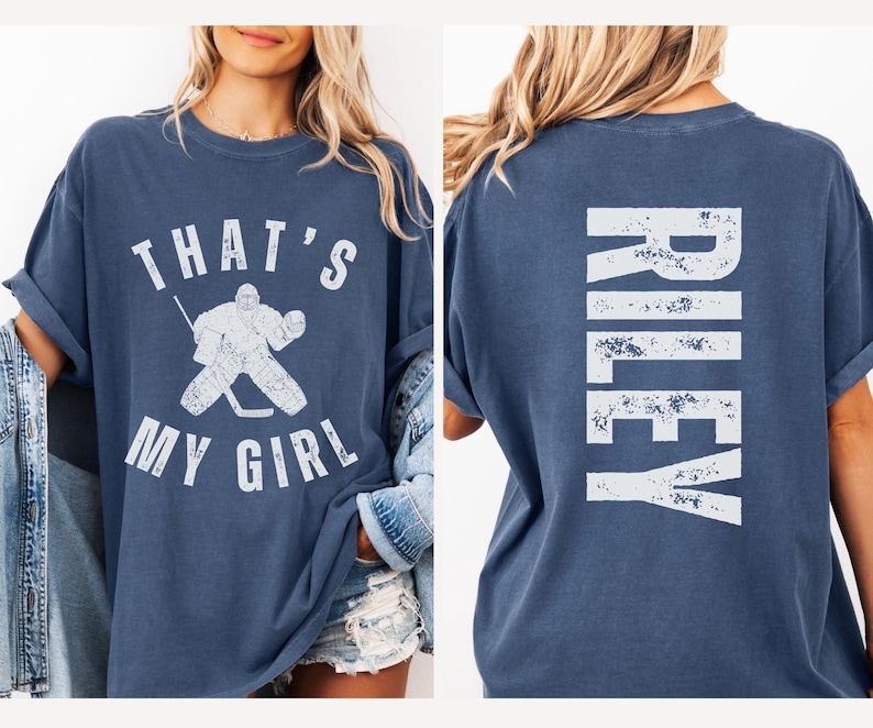 May include: A blue t-shirt with a white graphic of a hockey player and the text "That's My Girl". The back of the shirt has the name "Riley" in white distressed lettering.