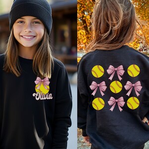 May include: Black sweatshirt with a softball and bow design on the front and multiple softball and bow designs on the back. The text "Olivia" is on the front of the sweatshirt.