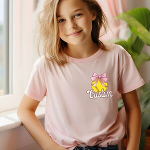 May include: A pink t-shirt with a yellow softball with a pink bow on top. The text "Custom" is written below the softball.