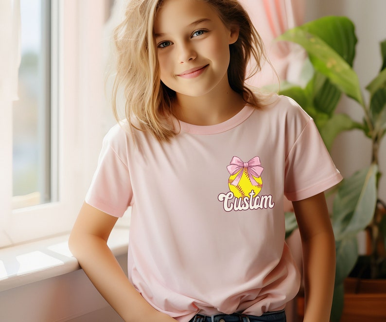 May include: A pink t-shirt with a yellow softball with a pink bow on top. The text "Custom" is written below the softball.