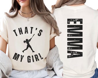 May include: A cream-colored t-shirt featuring a black graphic on the front with the text THAT'S MY GIRL and a silhouette of a softball player swinging a bat. The back of the shirt displays the name EMMA in large, vertical, distressed black letters.