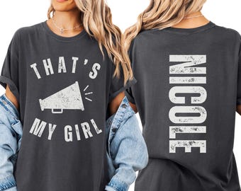 May include: A charcoal gray short-sleeve t-shirt featuring a white distressed graphic of a megaphone with the text THAT'S MY GIRL on the front and the name NICOLE printed vertically in white distressed lettering on the back.