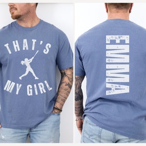 May include: Two blue t-shirts with white text. One shirt says "That's My Girl" with a silhouette of a woman playing baseball. The other shirt says "Emma" vertically down the back.