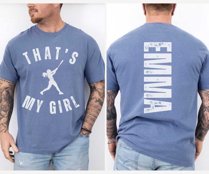 May include: Two blue t-shirts with white text. One shirt says "That's My Girl" with a silhouette of a woman playing baseball. The other shirt says "Emma" vertically down the back.