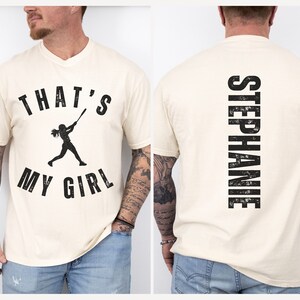 May include: A cream colored t-shirt with a black graphic of a girl playing baseball and the text "That's My Girl". The back of the shirt has the text "Stephanie" in black.