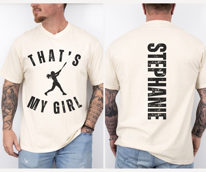 May include: A cream colored t-shirt with a black graphic of a girl playing baseball and the text "That's My Girl". The back of the shirt has the text "Stephanie" in black.