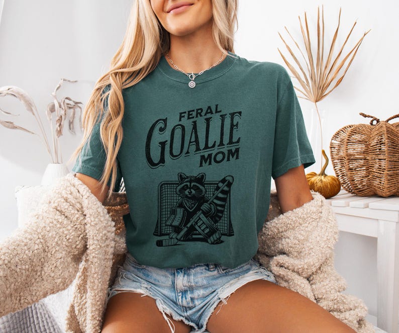 May include: A teal blue t-shirt with a black graphic of a raccoon wearing a goalie mask and holding a hockey stick. The text "Feral Goalie Mom" is printed on the shirt.