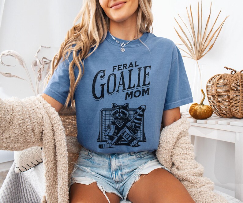 May include: A woman wearing a blue t-shirt with the text "Feral Goalie Mom" and a graphic of a raccoon in a hockey goalie outfit.