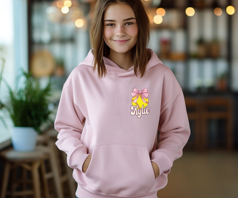 May include: A pink hoodie with a yellow softball with a pink bow and the name "Kylie" printed on it.