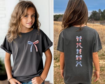 May include: Dark gray t-shirt featuring a patriotic bow design on the front and back. The front bow is blue, red, and white. The back of the shirt has four bows in a vertical row. The shirt is worn by a young person.