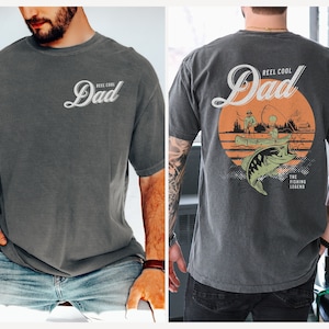 May include: A dark gray t-shirt with the text "Reel Cool Dad" and a graphic of a fishing scene with a sunset and a fish. The text "The Fishing Legend" is also on the shirt.