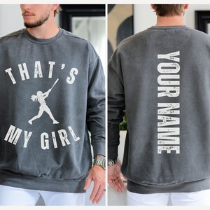 May include: A gray sweatshirt with a white graphic on the front that says "That's My Girl" with a silhouette of a girl playing baseball. The back of the sweatshirt has the text "YOUR NAME" in white.