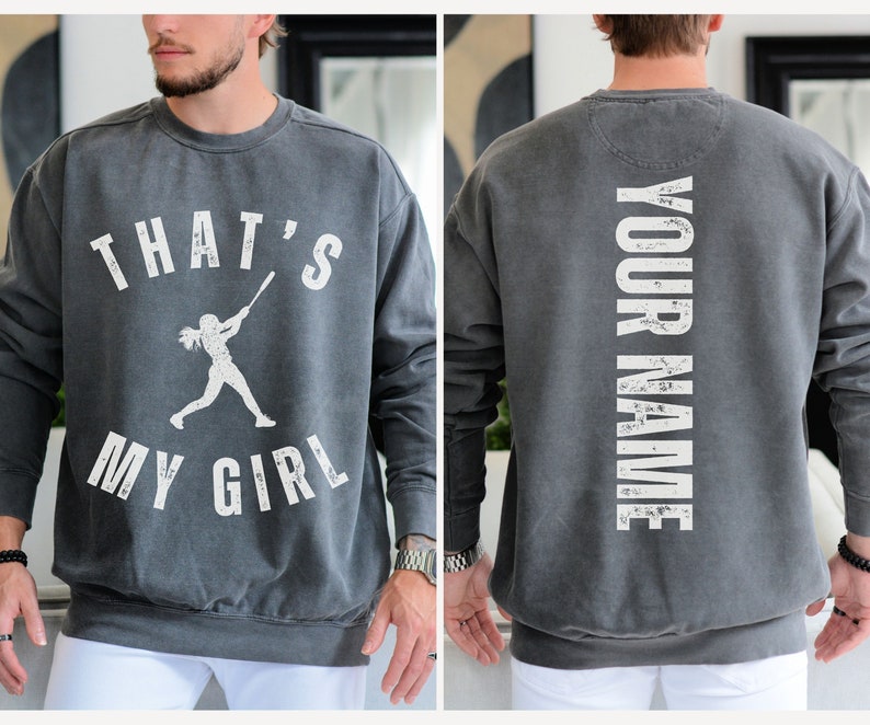 May include: A gray sweatshirt with a white graphic on the front that says "That's My Girl" with a silhouette of a girl playing baseball. The back of the sweatshirt has the text "YOUR NAME" in white.