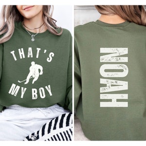 Może przedstawiać: Zielony sweter z napisem "That's My Boy" i grafiką hokeisty z przodu. Z tyłu swetra znajduje się napis "Noah" w białym kolorze.
