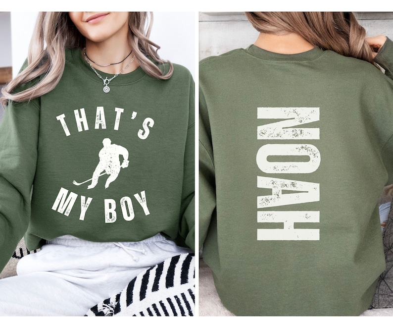 Może przedstawiać: Zielony sweter z napisem "That's My Boy" i grafiką hokeisty z przodu. Z tyłu swetra znajduje się napis "Noah" w białym kolorze.