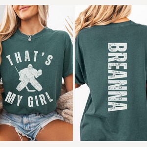 May include: A teal green t-shirt with a white graphic on the front that says "That's My Girl" with a hockey player illustration. The back of the shirt has the word "Breanna" in white.