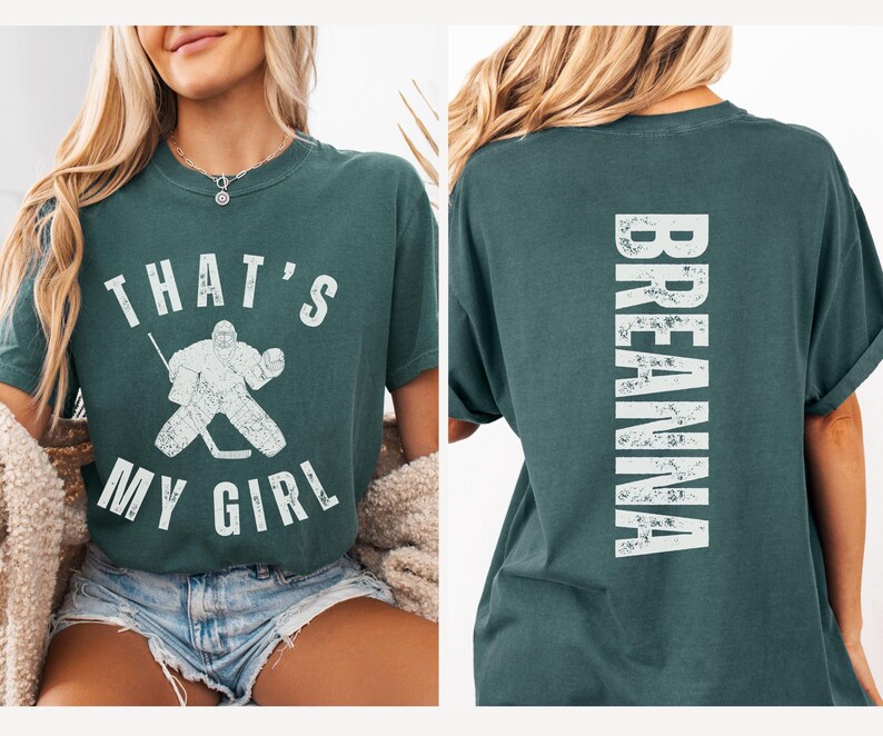 May include: A teal green t-shirt with a white graphic on the front that says "That's My Girl" with a hockey player illustration. The back of the shirt has the word "Breanna" in white.