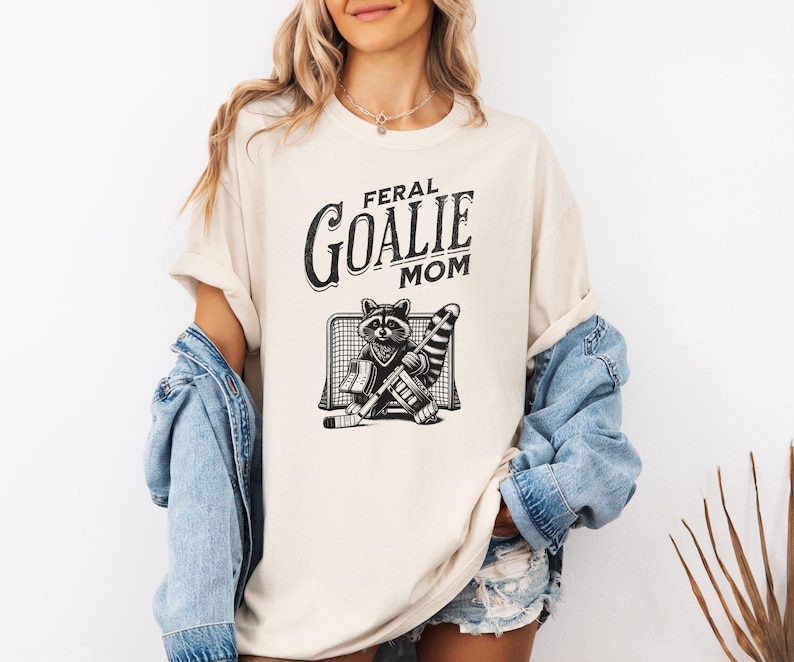 May include: A cream-colored t-shirt with the text "Feral Goalie Mom" in black lettering. A black and white illustration of a raccoon wearing hockey gear and standing in front of a hockey net is below the text.