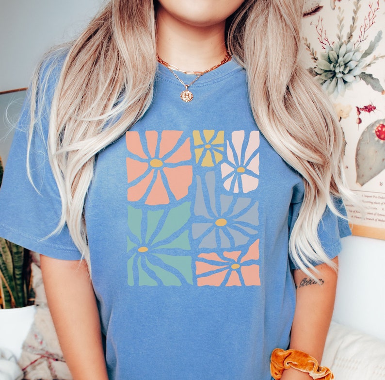 Op de afbeelding: Een lichtblauw T-shirt met een kleurrijk bloemenontwerp. Het ontwerp bevat een verscheidenheid aan bloemen in tinten roze, blauw, groen en geel.