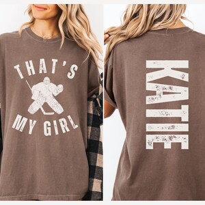 May include: A brown t-shirt with a white graphic of a hockey goalie and the text "That's My Girl". The back of the shirt has the name "Katie" in white text.